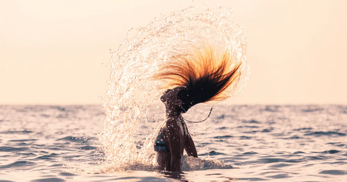 Donna in mare che getta indietro i capelli bagnati sollevando un ventaglio di gocce, al tramonto